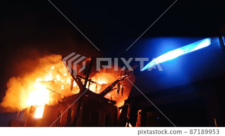 The roof of the house is on fire. The residential building burn, village. Firefighters put out a fire. Fire truck, headlights and panel, siren signal. 87189953