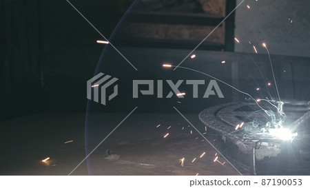 Forge workshop. Smithy. Worker in a welding hood helmet welds a part by electric welding. Sparks are reflected in the protective screen. Forge workshop. Smithy. Worker in a welding hood helmet welds a part by electric welding. Sparks are reflected in the protective screen. 87190053