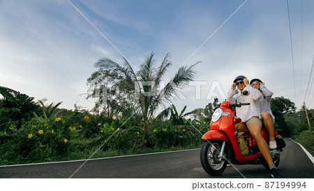 Love couple on red motorbike in white clothes to go on forest road trail trip. Two caucasian tourist woman man drive on scooter. Motorcycle rent. 87194994