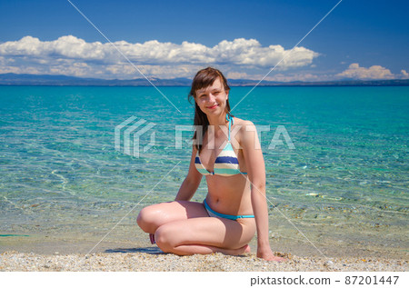 Young beautiful girl in bikini sit on sandy beach near turquoise water of Toroneos kolpos gulf 87201447