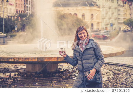 Young woman tourist with grey jacket smile and hold therapeutic mineral water Young woman tourist with grey jacket smile and hold therapeutic mineral water 87201595