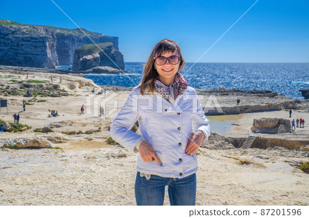 Young woman traveler with white jacket and sunglasses looking at camera and smile on coast of Dwejra Bay 87201596