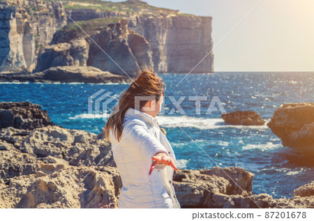 Young woman traveler with white jacket and headscarf in the wind looking away distance on stone rocks of Dwejra Bay 87201678