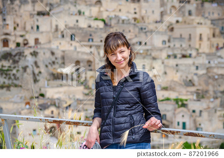 Young beautiful woman traveler with black jacket looking at camera, smile, posing and keep spikelet in Sassi di Matera Young beautiful woman traveler with black jacket looking at camera, smile, posing and keep spikelet in Sassi di Matera 87201966