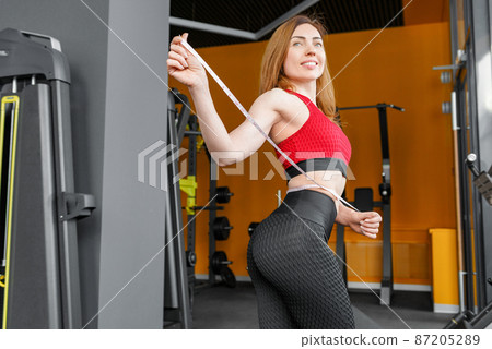 Fit woman refreshing after workout. Close up of slim woman measuring her waists size with tape measure. 87205289