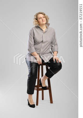 Senior woman in grey shirt and faux leather trousers sits on tall wooden chair over grey studio background 87219551