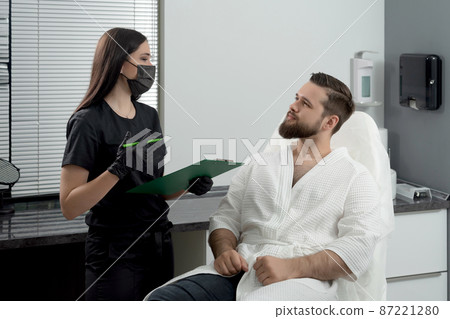 Expert female cosmetologist giving consultation to young man patient sitting on a couch in beautician office of aesthetic clinic 87221280