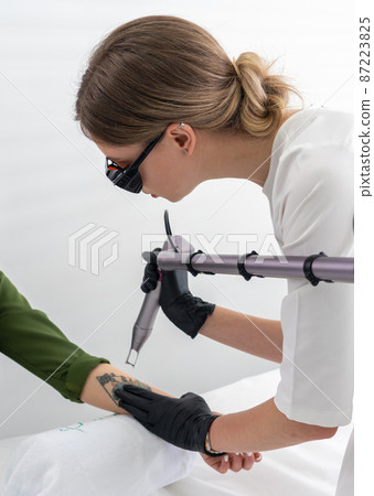 Cosmetologist using laser device to remove an unwanted tattoo from female arm. Concept of erasing tattoos as an expensive procedure in a cosmetology clinic 87223825