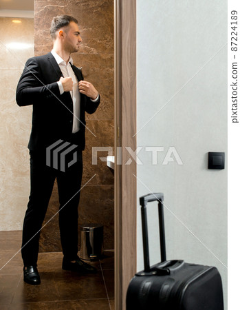 Young traveler businessman wearing white shirt standing at the mirror and getting dressed for meeting or work after arriving in the hotel room with his luggage Young traveler businessman wearing white shirt standing at the mirror and getting dressed for meeting or work after arriving in the hotel room with his luggage 87224189