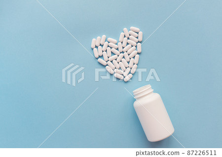 White pills in the shape of a heart and a white jar on a blue background. Pills for heart health. The concept of medicine and health care. 87226511