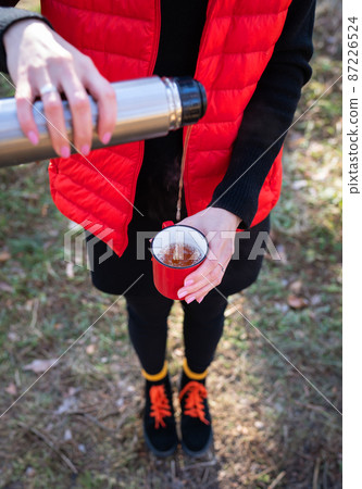 A girl in a bright orange vest pours hot tea from a thermos, a snack in the open air.Travel, trekking and hiking. A girl in a bright orange vest pours hot tea from a thermos, a snack in the open air.Travel, trekking and hiking. 87226524