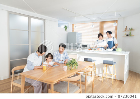 A young couple cooking in the kitchen with their grandparents watching a tablet with an elementary school girl in the dining room, three generations 87230041