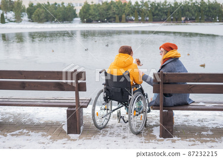 Caucasian woman in a wheelchair and her friend are sitting by the lake with ducks in winter. Caucasian woman in a wheelchair and her friend are sitting by the lake with ducks in winter. 87232215
