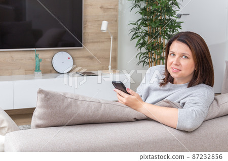 Young woman looking at her smart phone and smiling while sitting on the couch Young woman looking at her smart phone and smiling while sitting on the couch 87232856