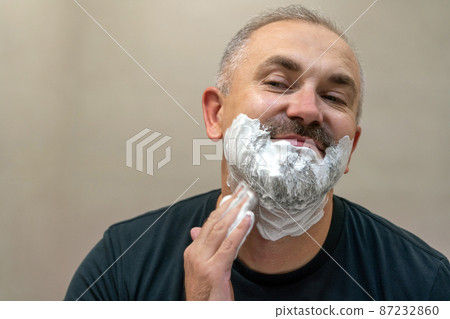 Portrait of handsome white-haired beared middle-aged man applying shaving foam to trim his beard Portrait of handsome white-haired beared middle-aged man applying shaving foam to trim his beard 87232860