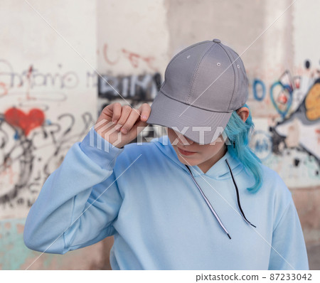 Blue haired Teenage girl in blue hoodie and baseball cap staying against graffiti wall 87233042