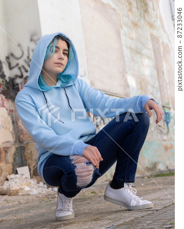 Blue haired Teenage girl in light blue oversize hoodie sitting on her haunches near graffiti wall 87233046