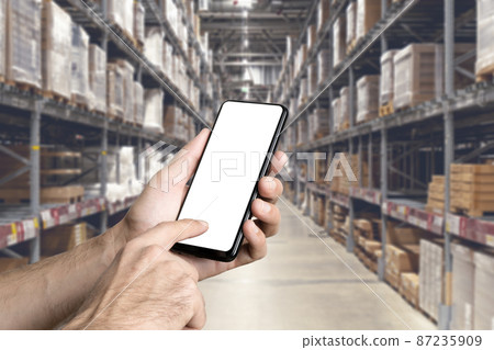 Warehouse with a hand holding an empty smartphone. Blurred industrial warehouse with goods and products. Logistics, business, delivery, services concept Warehouse with a hand holding an empty smartphone. Blurred industrial warehouse with goods and products. Logistics, business, delivery, services concept 87235909