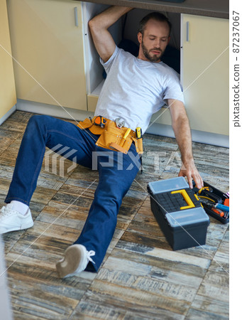 Plumbing expert. Professional repairman, plumber wearing tool belt examining and fixing sink pipe in the kitchen indoors 87237067