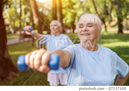 Strong senior woman does exercises with dumbbell training with husband in green park Strong senior woman does exercises with dumbbell training with husband in green park 87237108
