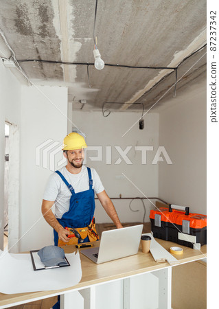 Happy repairman worker in uniform wearing tool belt and hardhat holding hammer and smiling at camera while standing indoors Happy repairman worker in uniform wearing tool belt and hardhat holding hammer and smiling at camera while standing indoors 87237342