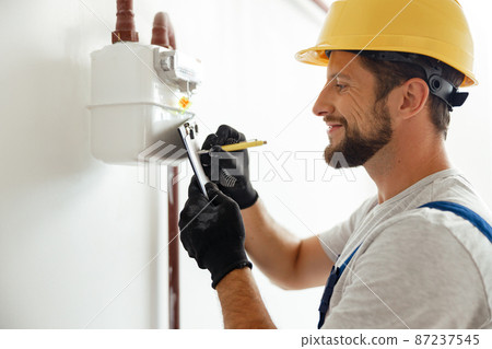 Happy professional heating fitter making notes while checking gas consumption at gas pipe and meter counter 87237545