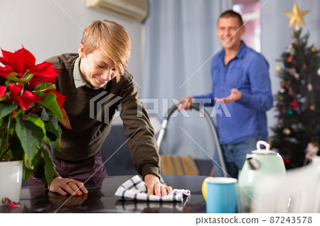 Son cleans the table with a rag while father vacuums room before christmas Son cleans the table with a rag while father vacuums room before christmas 87243578