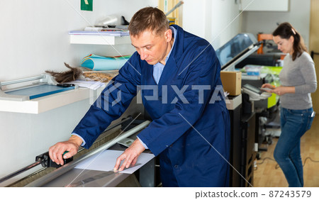 Printing worker cuts paper on a cutter machine Printing worker cuts paper on a cutter machine 87243579