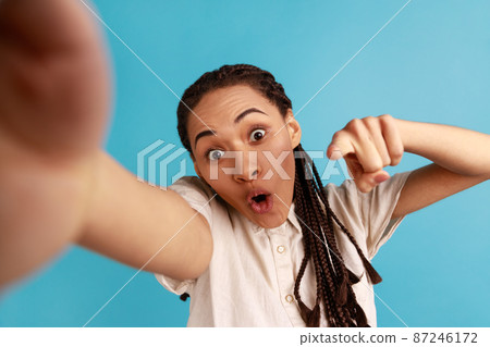 Shocked woman with black dreadlocks has astonished face expression, making selfie, expresses surprised emotions, pointing finger to camera, POV. Indoor studio shot isolated on blue background. Shocked woman with black dreadlocks has astonished face expression, making selfie, expresses surprised emotions, pointing finger to camera, POV. Indoor studio shot isolated on blue background. 87246172