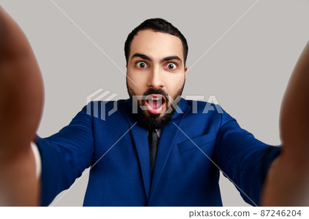 Astonished bearded young adult man taking selfie, looking at camera with shocked expression and open mouth POV, wearing official style suit. Indoor studio shot isolated on gray background. 87246204