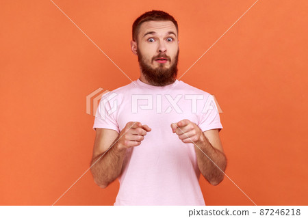 Portrait of shocked surprised bearded man looking with big eyes and open mouth, pointing finger at camera, suspecting you, wearing pink T-shirt. Indoor studio shot isolated on orange background. Portrait of shocked surprised bearded man looking with big eyes and open mouth, pointing finger at camera, suspecting you, wearing pink T-shirt. Indoor studio shot isolated on orange background. 87246218