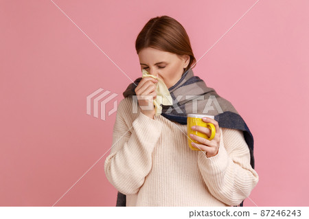 Portrait of unhealthy sick ill blond woman wrapped in scarf standing with cup of tea, suffering from runny nose, wearing white sweater. Indoor studio shot isolated on pink background. Portrait of unhealthy sick ill blond woman wrapped in scarf standing with cup of tea, suffering from runny nose, wearing white sweater. Indoor studio shot isolated on pink background. 87246243