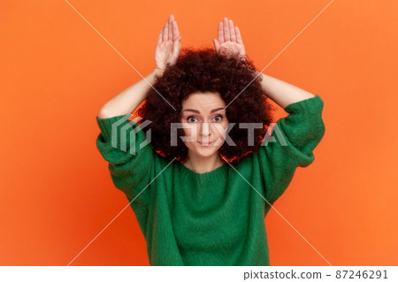 Woman with Afro hairstyle wearing green casual style sweater showing bunny ears gesture, holding hands on head and smiling at camera, having fun. Indoor studio shot isolated on orange background. Woman with Afro hairstyle wearing green casual style sweater showing bunny ears gesture, holding hands on head and smiling at camera, having fun. Indoor studio shot isolated on orange background. 87246291