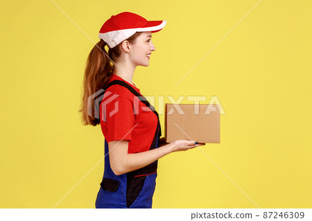 Side view of delighted courier woman standing with cardboard parcel, giving it to client, looking at camera, wearing overalls and red cap. Indoor studio shot isolated on yellow background. Side view of delighted courier woman standing with cardboard parcel, giving it to client, looking at camera, wearing overalls and red cap. Indoor studio shot isolated on yellow background. 87246309