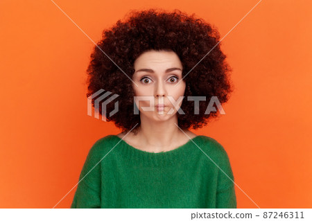 Funny woman with Afro hairstyle in green sweater making fish face with pout lips and looking amazed, choking with idiotic comical silly expression. Indoor studio shot isolated on orange background. Funny woman with Afro hairstyle in green sweater making fish face with pout lips and looking amazed, choking with idiotic comical silly expression. Indoor studio shot isolated on orange background. 87246311