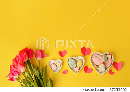 Sweets, pastry, gingerbread cookies for Easter table. Easter eggs heart shaped decor plate, pink tulips on yellow background top view copy space, spring seasonal holiday banner for site, flyer, coupon 87253302