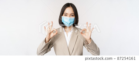 Coronavirus and workplace concept. Image of asian saleswoman, company worker in medical mask showing okay sign, smiling pleased, white background 87254161