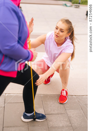 Vertical cropped shot obese overweight young woman doing exercises using fitness tape for weight loss with personal trainer outdoor in summer day. Fat female with coach stretching before running. Vertical cropped shot obese overweight young woman doing exercises using fitness tape for weight loss with personal trainer outdoor in summer day. Fat female with coach stretching before running. 87254666