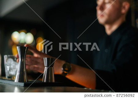 Low-angle view of professional bartender preparing refreshing alcoholic cocktail standing behind bar counter, focus in foreground to metal jigger, in modern nightclub with dark interior. 87256092