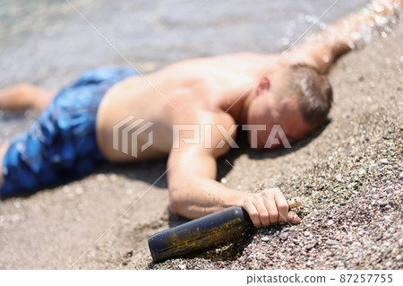 A drunk man with a bottle lies on the wet sand, blurry 87257755