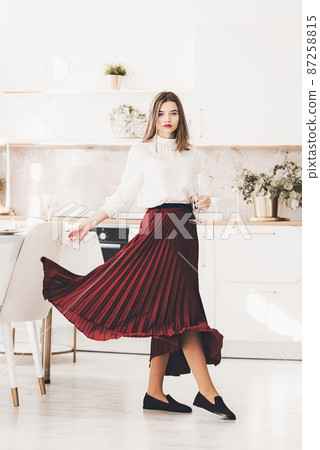 Portrait of fashionable woman in a red skirt, white blouse and stylish suede shoes with a buckle posing on the kitchen. Girl with a vine glass in a hands Portrait of fashionable woman in a red skirt, white blouse and stylish suede shoes with a buckle posing on the kitchen. Girl with a vine glass in a hands 87258815