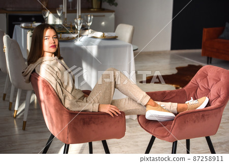 photo of a women in beige sports suit and stylish leather sneakers posing in the chair on a kitchen . selective focus photo of a women in beige sports suit and stylish leather sneakers posing in the chair on a kitchen . selective focus 87258911