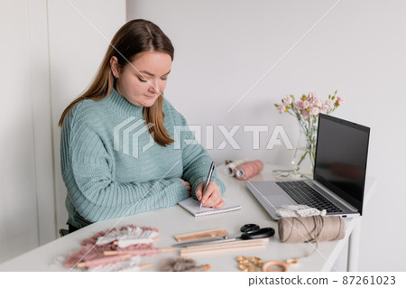 Young stylish woman using laptop at home, writing to do list. Female hobby. Working on research project online concept. Handmade macrame shop 87261023