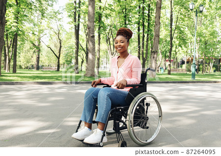 Beautiful young black woman in wheelchair going for walk at city park, posing and smiling outdoors, free space 87264095