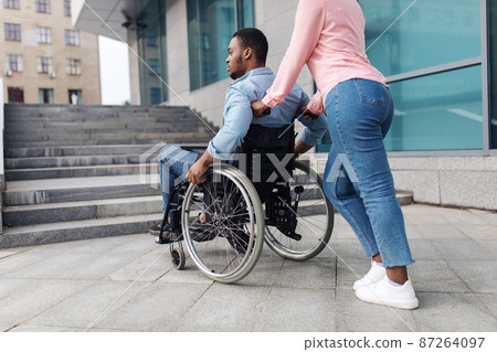 Young black woman with millennial man in wheelchair having no possibility to enter building without ramp, outdoors Young black woman with millennial man in wheelchair having no possibility to enter building without ramp, outdoors 87264097