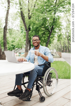 Happy young black man in wheelchair using laptop and drinking coffee at table in outdoor cafe 87264184