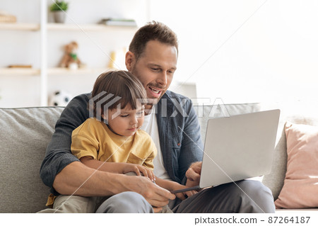 Family business concept. Young father businessman working on laptop at home, showing his son documents and explaining Family business concept. Young father businessman working on laptop at home, showing his son documents and explaining 87264187