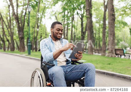 Happy young black man in wheelchair using tablet pc, browsing web or watching movie at spring park Happy young black man in wheelchair using tablet pc, browsing web or watching movie at spring park 87264188