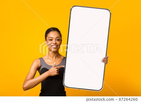 Smiling young slim black female points finger at huge smartphone with empty screen, isolated on orange background Smiling young slim black female points finger at huge smartphone with empty screen, isolated on orange background 87264260