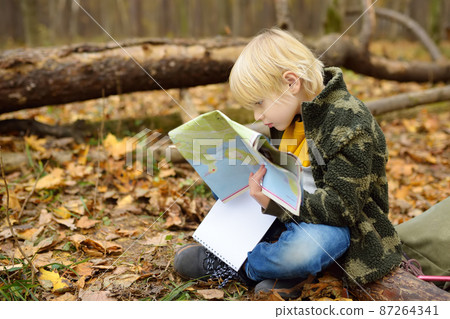 Little boy scout is orienteering in forest. Child is sitting on fallen tree and looking on map on background of teepee hut. Adventure, scouting and hiking tourism for kids. Little boy scout is orienteering in forest. Child is sitting on fallen tree and looking on map on background of teepee hut. Adventure, scouting and hiking tourism for kids. 87264341